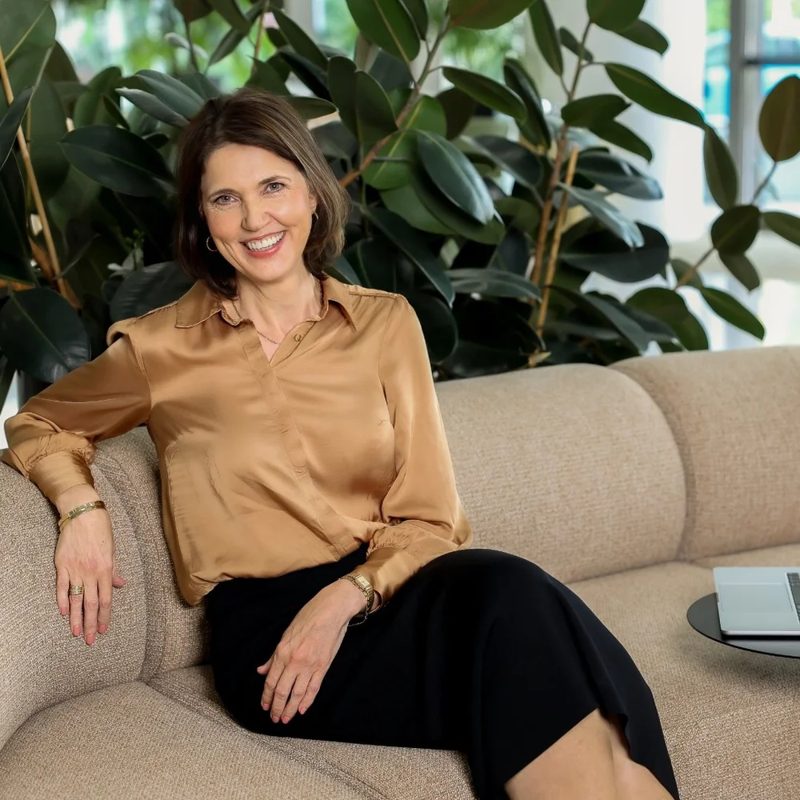 Annelie Peusschers, smiling, seated on a sofa in front of large green plants
